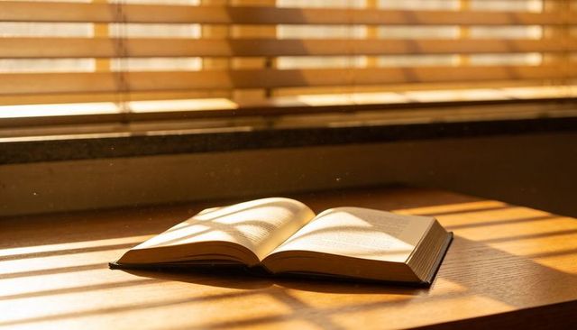 Sunlit open hardcover book on wooden table with blinds shadows and floating dust