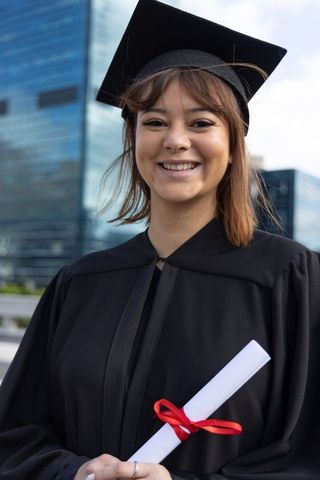 Smiling asian graduate woman wearing cap and gown holding diploma in urban campus