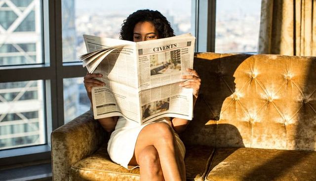 Woman reading newspaper on tufted gold sofa by city window in sunlit luxury apartment