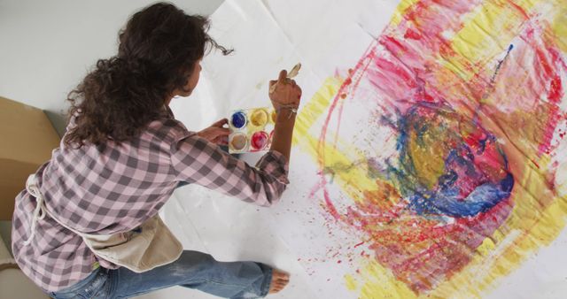 Woman creating large abstract painting with bright colors on canvas. Visible paint splashes and brush strokes add dynamic movement. Great for promoting artistic workshops, creativity, art classes, studio environments, and inspired living.