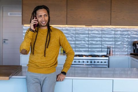 African american man leaning on kitchen island speaking on smartphone while working remotely