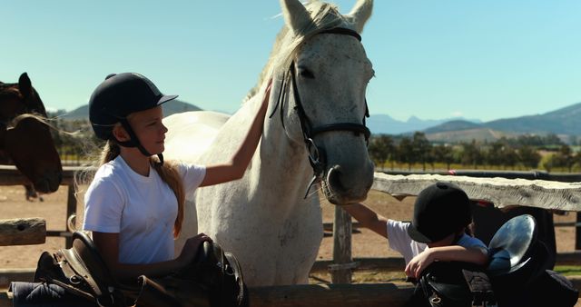 Children preparing their horses for a ride on a sunny day at a ranch. The kids wearing helmets interact with horses, emphasizing animal care and outdoor activities. Suitable for promoting equestrian activities, ranch visits, and outdoor educational programs.