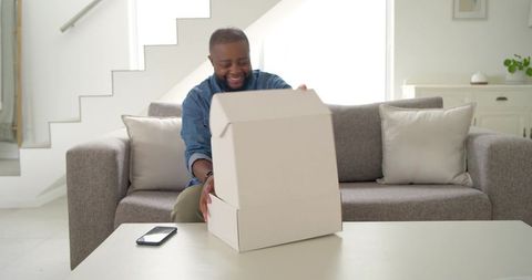 African american man opening delivery box on sofa in bright modern minimalist living room