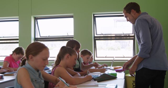 Teacher engaging with group of students in a brightly lit classroom with green walls. Students are focused on different activities, such as writing and using educational tools. Ideal for illustrating educational environments, teaching methods, and primary school learning. Can be used in materials related to education, school brochures, teacher training, and child development studies.
