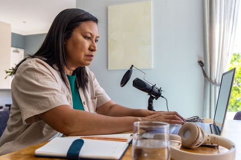 Mature woman recording podcast from home while typing on laptop with microphone
