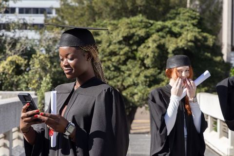 Graduating students celebrating in caps and gowns checking phone and holding diplomas