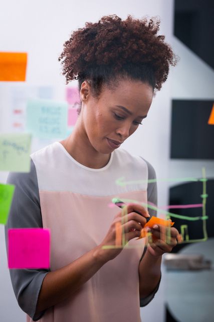 Businesswoman brainstorming and writing ideas on sticky notes attached to a glass wall in an office. Ideal for use in articles or presentations about creative business strategies, planning sessions, professional work environments, and innovative thinking.