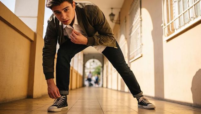 Leaning forward young man adjusting sneaker on tiled corridor in sunlit urban arcade