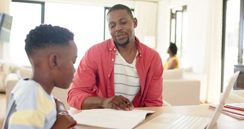 Father helping son with homework using laptop and workbook in bright living room