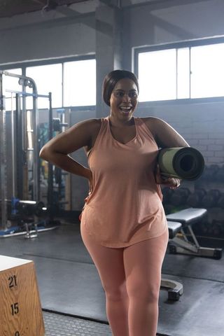 Confident african american woman carrying yoga mat in sunlit industrial gym wearing peach activewear