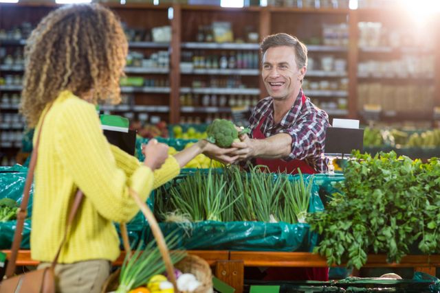 Male staff member in a supermarket smiling and assisting a woman with her grocery shopping. The woman is holding a basket filled with fresh produce. Ideal for use in articles or advertisements related to customer service, grocery shopping, healthy eating, and retail environments.