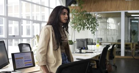Indian businesswoman leaning on chair working in modern open office wearing blazer and jeans