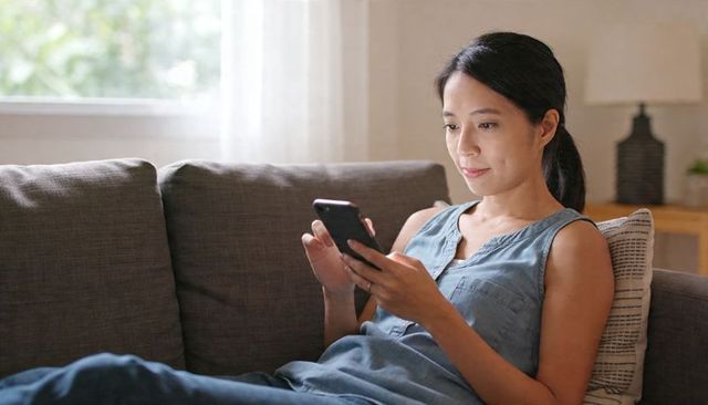 Reclining asian woman using smartphone on gray sofa in cozy minimalist living room, natural daylight