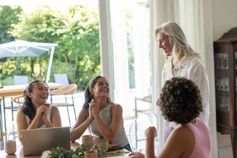 Intergenerational women laughing and chatting around sunlit dining table with laptop