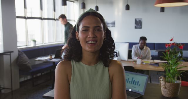 Smiling businesswoman stands in modern office space while colleagues collaborate in background. Plants and natural light create warm, inviting atmosphere. Perfect for themes of teamwork, productivity, modern work environments, and diversity.