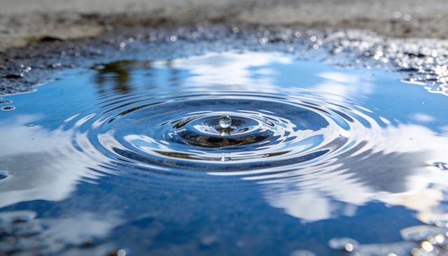 Water droplet creating concentric ripples in reflective puddle with sky reflection