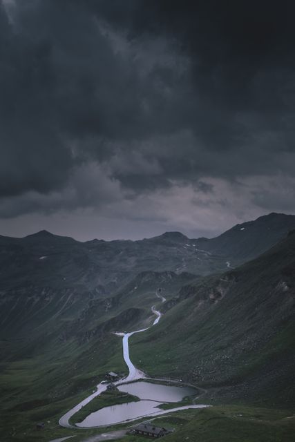A winding road cuts through a mountainous landscape under a dark, stormy sky. The dramatic clouds create a moody atmosphere at dusk, highlighting the rugged beauty of this alpine wilderness. This image can be used for travel and adventure blogs, nature magazines, or to evoke a sense of journey and exploration in advertising and promotional materials.