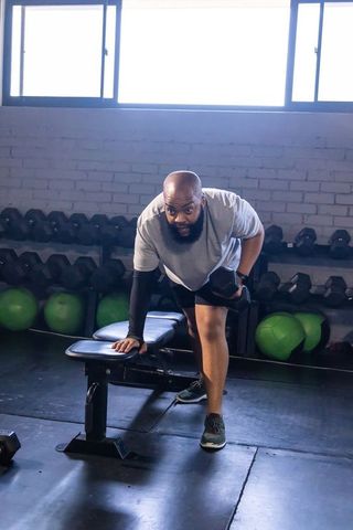 African american man performing one-arm hex dumbbell row on bench in gym natural light