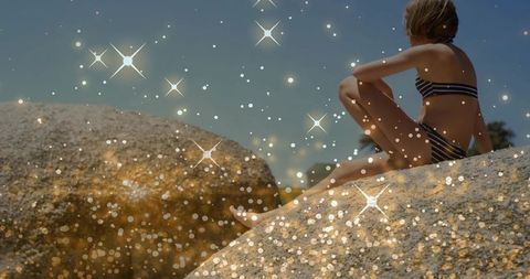 Child sitting on sunlit granite rocks wearing striped swimsuit gazing at sea with sparkles