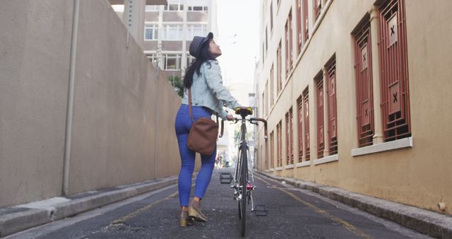 A woman is walking her bicycle down a narrow city alley. She is wearing a denim jacket, jeans, and a hat, carrying a brown bag. This image is perfect for depicting urban lifestyle, transportation themes, or modern city fashion. It can be used for blogs, marketing materials, or magazines focusing on urban living, style, or transportation.