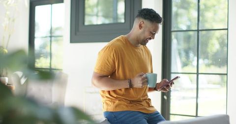 Sunlit home moment: african american man smiling at phone while holding coffee mug