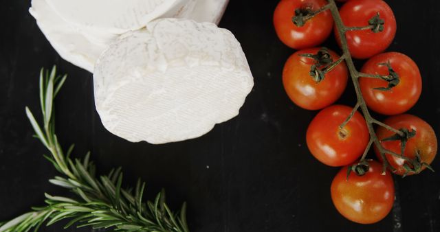 A wheel of Brie cheese is paired with a sprig of rosemary and a vine of cherry tomatoes on a dark surface, with copy space. Fresh ingredients like these are often used to create flavorful and visually appealing culinary presentations.