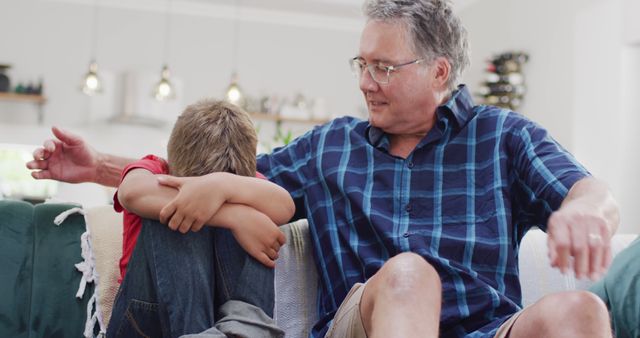 Mature man with gray hair and glasses comforting his young grandson who appears sad while sitting on a couch in a cozy living room. This image conveys themes of family, care, and emotional support. Useful for content on family relationships, elder care, emotional well-being, and parenting.