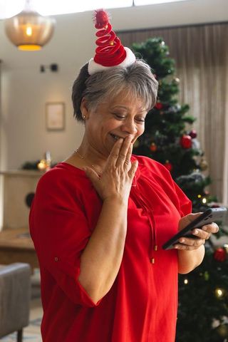 Elderly woman joyfully using smartphone by christmas tree at home