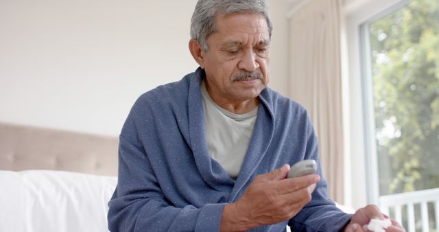 Senior biracial man in bathrobe checking blood sugar level with glucometer at home. Healthcare, retirement, wellness and senior lifestyle, unaltered.