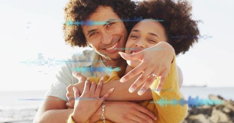 Couple embracing on rocky shore showing engagement ring with waveform overlay, smiling