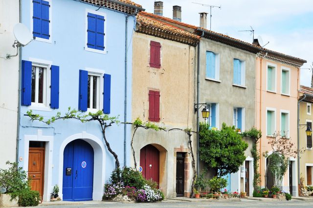 Image shows a row of colorful houses in a European village, each with brightly colored shutters and doors. This scenic view features houses painted in shades of blue, red, beige, and peach, and is a perfect representation of traditional European architecture. The street is lined with potted flowers and creeping ivy. This image is ideal for uses in travel blogs, tourism promotional materials, real estate, architectural studies, and cultural articles.