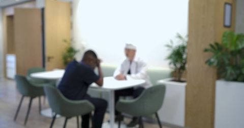 Clinician consulting patient at bright clinic table with potted plants and chairs in lounge