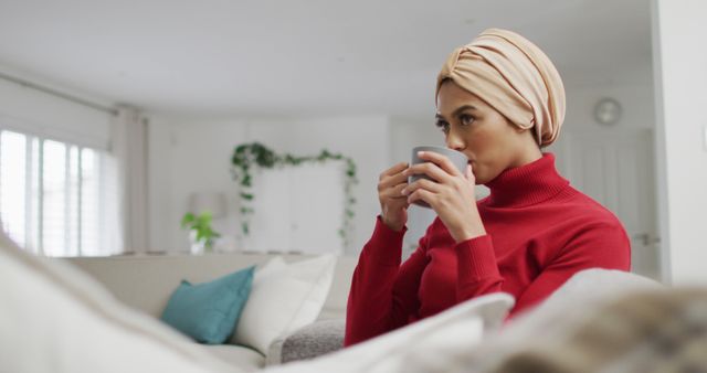 Image of biracial woman in hijab sitting on sofa and drinking coffee. Lifestyle, spending free time at home concept.