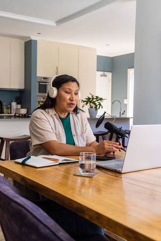 Mature woman podcasting at kitchen table wearing headphones recording with laptop and microphone