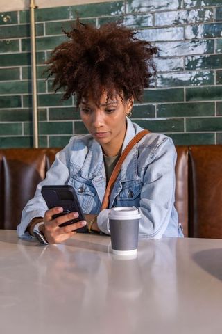 African american woman checking smartphone in urban cafe booth with takeaway coffee