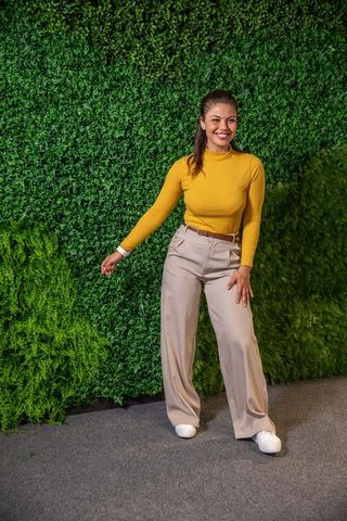 Smiling woman posing in mustard knit top and beige wide-leg trousers against lush green wall