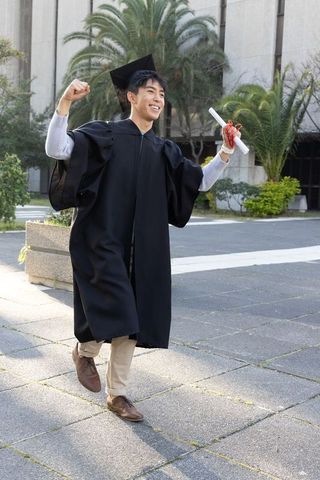 Triumphant asian graduate striding on campus in cap and gown holding diploma celebrating success