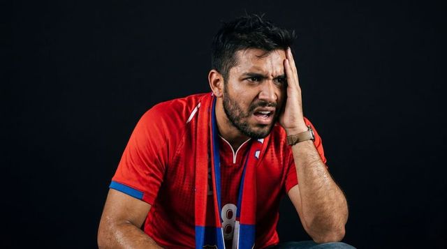 Indian sports fan grimacing while pressing hand to head wearing red jersey 8 and scarf