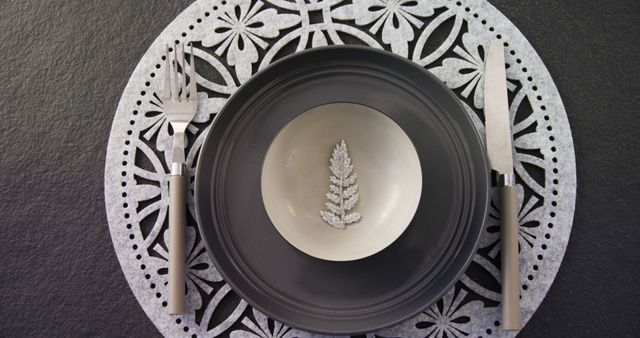 A decorative leaf motif adorns the center of a minimalist place setting featuring a gray plate, complemented by a pair of silver forks on a lacy placemat. The elegant simplicity of the tableware suggests a modern dining aesthetic or a sophisticated event setting.