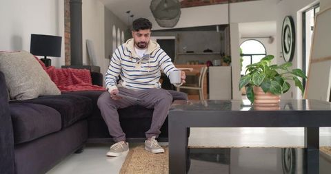 Indian man sipping coffee while using tablet on sectional sofa in minimalist living room