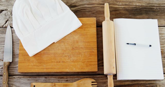 A chef's white hat and a rolling pin lie on a wooden surface next to a notebook and pen, with copy space. These items suggest a culinary setting, for recipe development or menu planning.