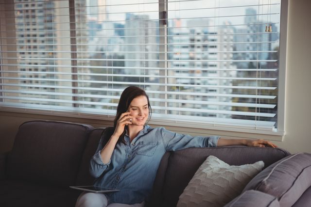 Woman sitting on a sofa in a modern living room, talking on a mobile phone. She is smiling and appears relaxed, suggesting a casual conversation. The background shows window blinds with a cityscape view, indicating an urban setting. This image can be used for themes related to communication, modern lifestyle, home comfort, and technology.