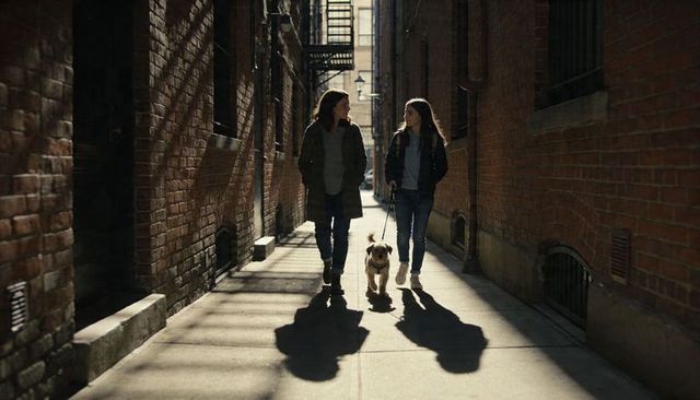 Mother and teen walking terrier down sunlit urban brick alley conversing casually
