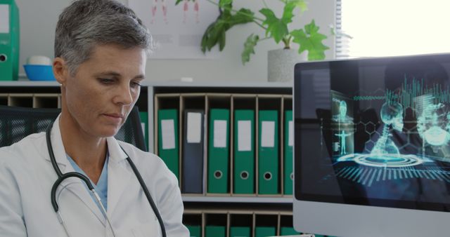 This image shows a female doctor in her office, focusing on a computer screen displaying medical technology visuals. She is wearing a stethoscope around her neck, suggesting that she is ready to attend to patients. This image is suitable for projects related to healthcare, medical research, patient care, technology in medicine, and professional medical settings.