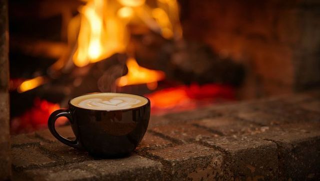 Steaming latte cup with heart foam resting on brick hearth by glowing fireplace, cozy warmth