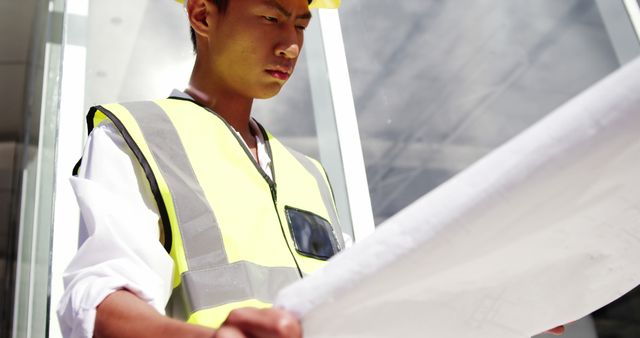 A young Asian construction worker in a yellow hard hat and reflective vest is intently reviewing a set of building plans, with copy space. His focused expression and the blueprint in his hands suggest he is carefully planning or inspecting a project.