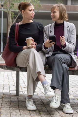 Female colleagues chatting on bench in urban plaza holding coffee and smartphone