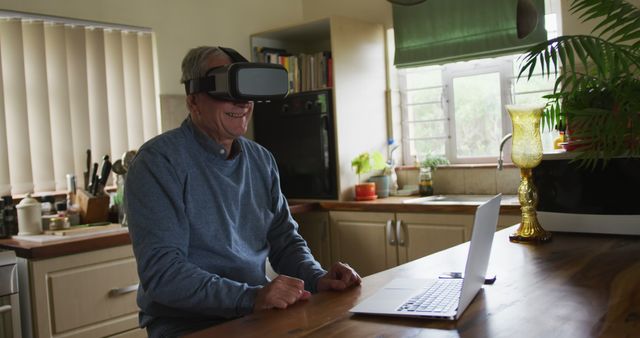 Senior man sits at wooden kitchen table, engaging with technology through a VR headset. The man is smiling, indicating enjoyment and a positive experience. Indoor environment with natural light coming through window. Great for illustrating themes of modern technology adoption by older generations, virtual interactions, and lifestyle. Suitable for promoting VR technology, senior lifestyle products, and home tech solutions.