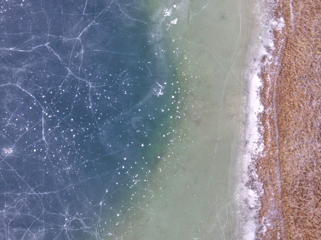 Aerial view of frozen lake shoreline showing cracked ice and snow. Perfect for illustrating winter landscapes, environmental themes, or geographical studies. Can be used in travel blogs, nature-focused articles, and educational content about winter and icy conditions.