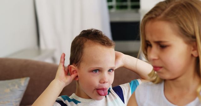 Boy making silly face with tongue out and hands over ears next to annoyed girl with blonde hair. Concept of playful interactions and family dynamics among young children. Ideal for advertisements, parenting articles, or family-focused campaigns.